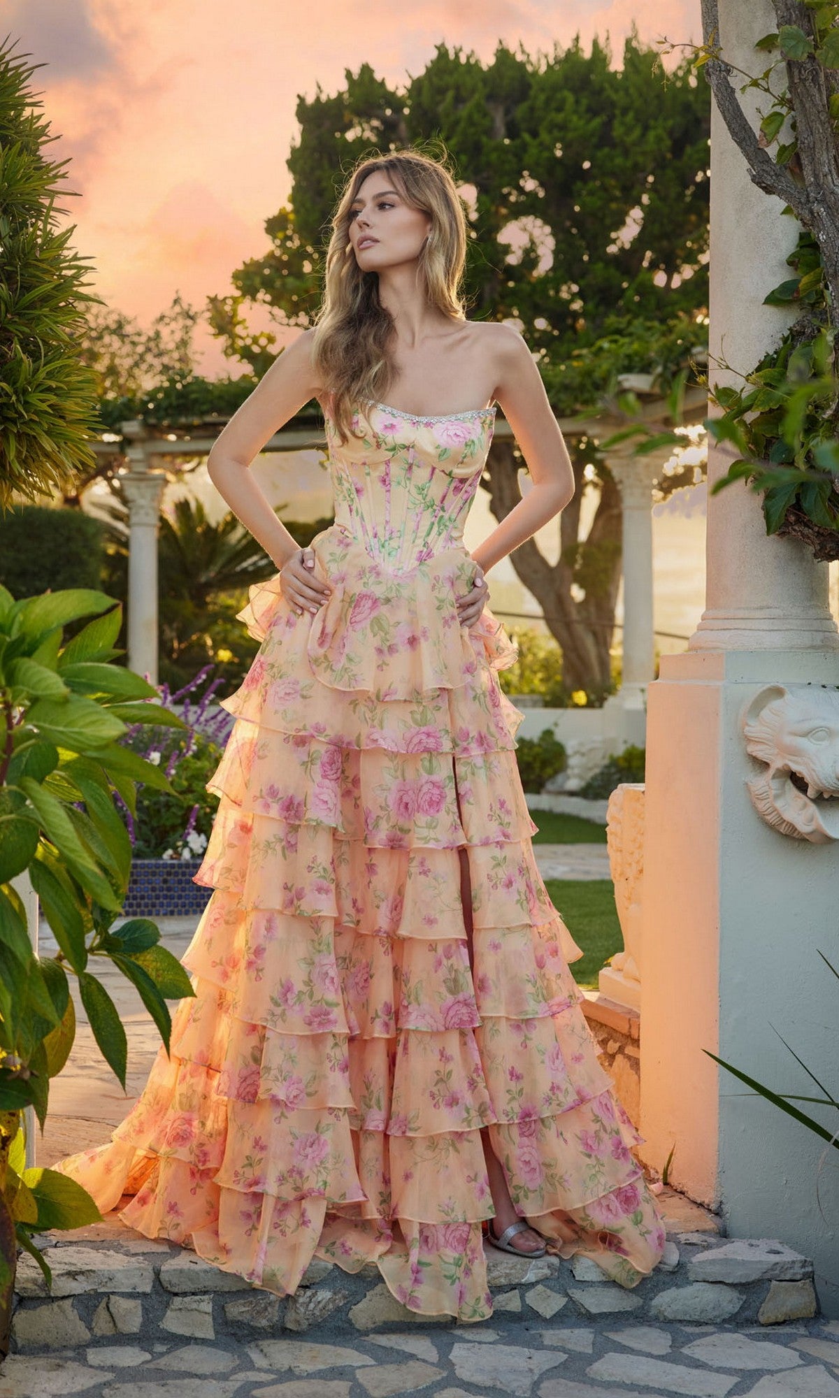 A woman stands outdoors at sunset before a garden pergola, wearing the Juliet JT2739S Long Prom Dress in Pastel Yellow Floral, featuring a strapless corset bodice and a flowing, ruffled chiffon A-line skirt with pink flowers.